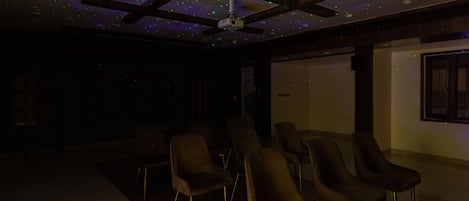 A dimly lit room with ceiling-mounted projectors and a ceiling fan, featuring rows of chairs and a projection screen.