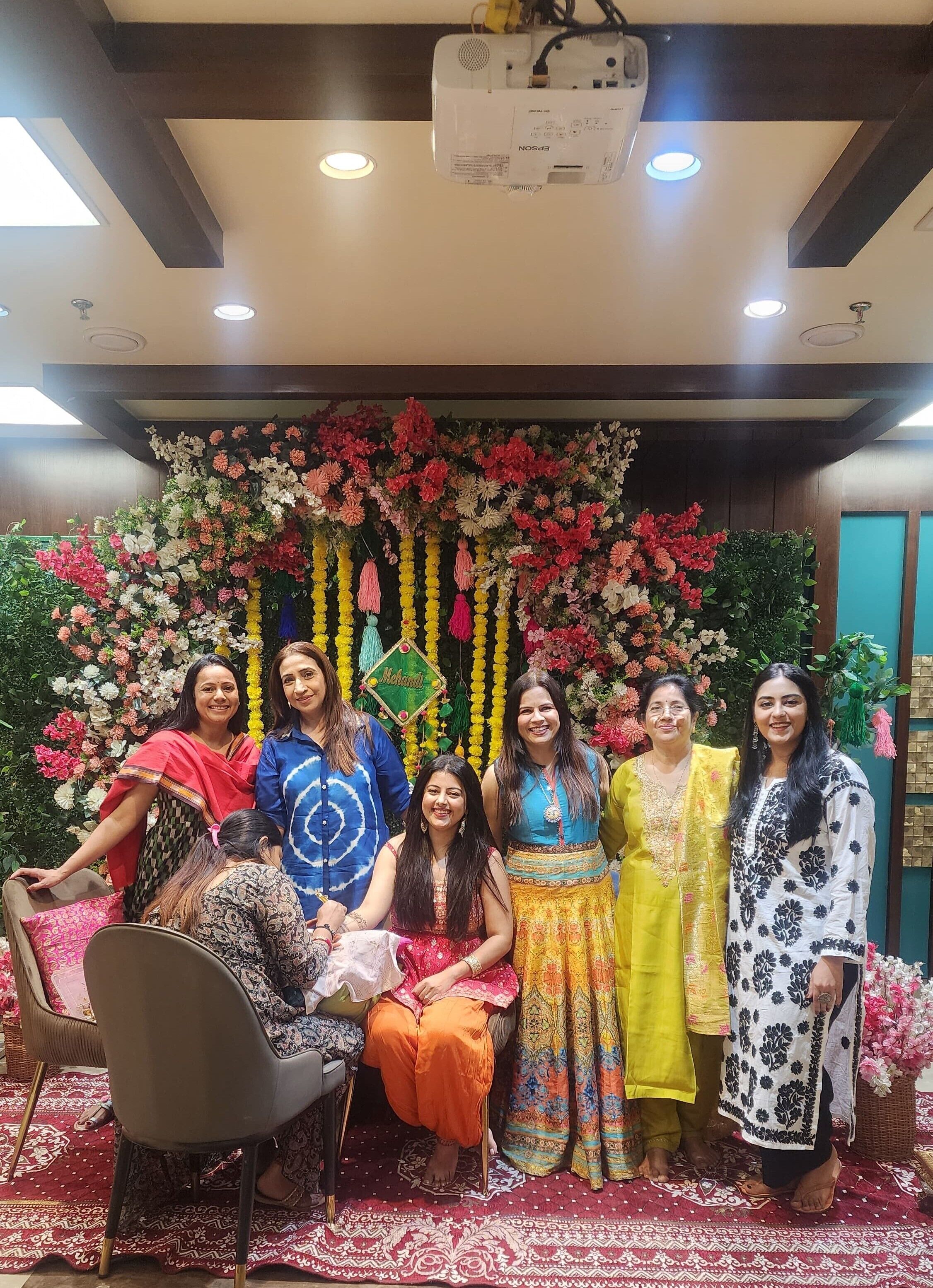Un grupo de personas sentadas y de pie en una habitación con un fondo decorativo de flores y luces en el techo.