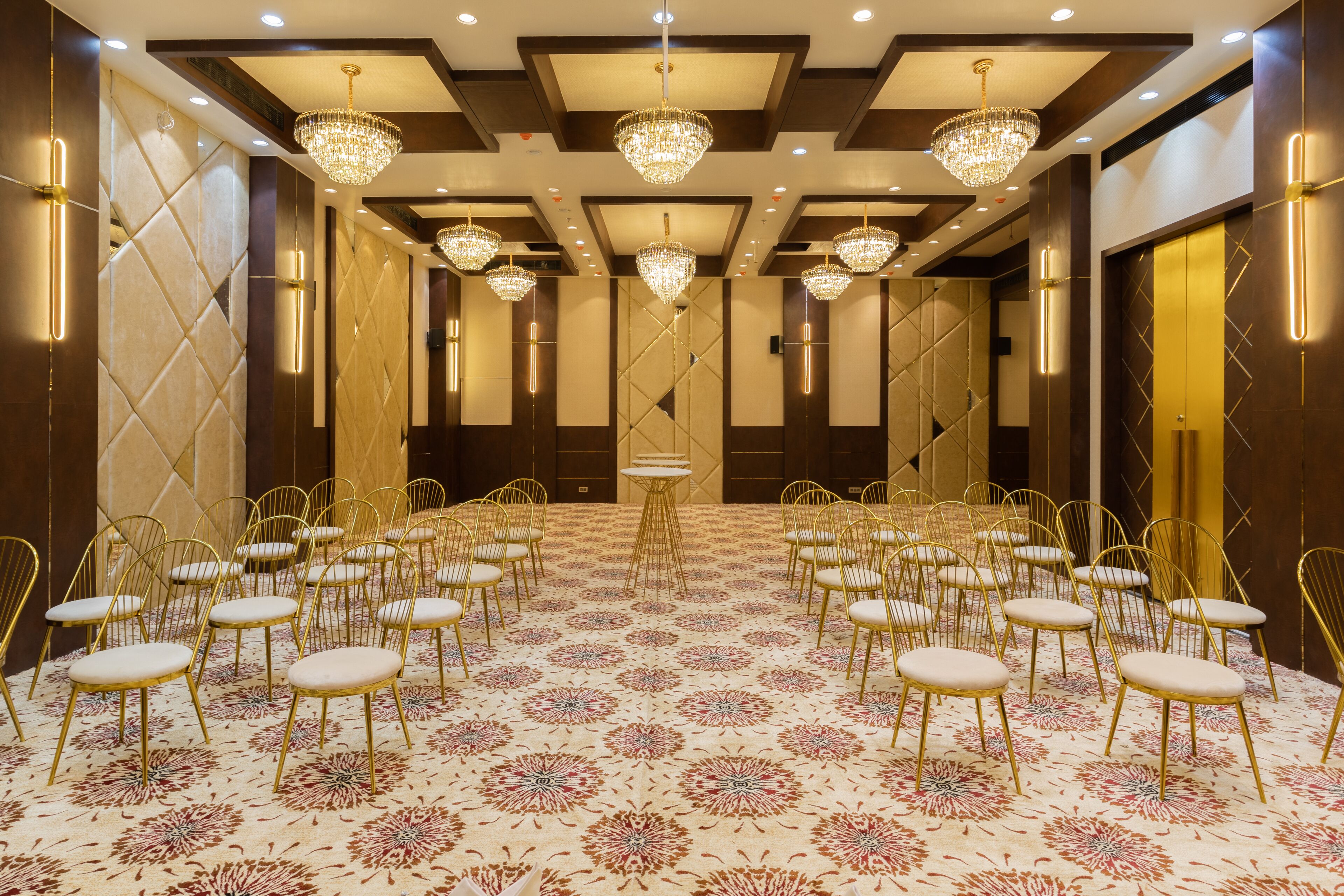 Un salón de conferencias con hileras de sillas doradas, alfombra estampada y varios candelabros.