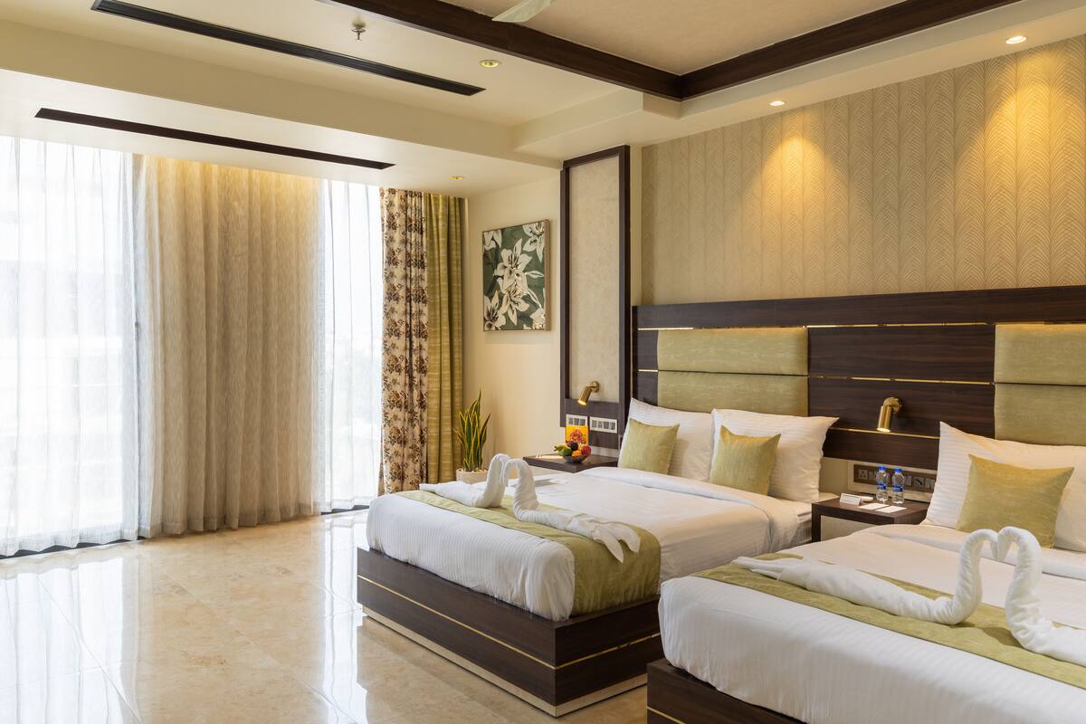 A hotel room with two beds, a large window with curtains, a wall-mounted artwork, and a nightstand with a lamp and water bottles.