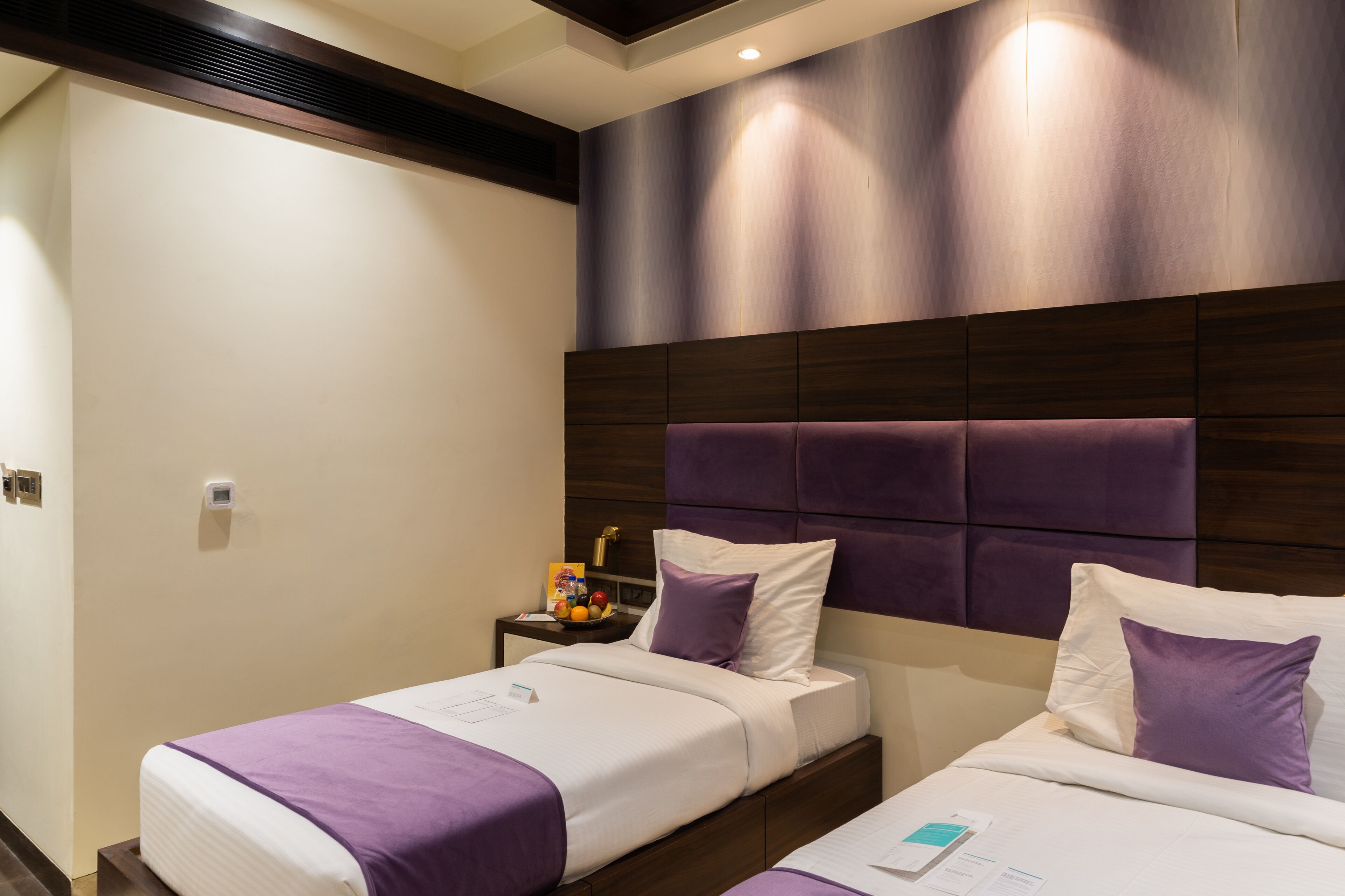 A hotel room with two beds, a wooden headboard, and a nightstand with a fruit bowl.