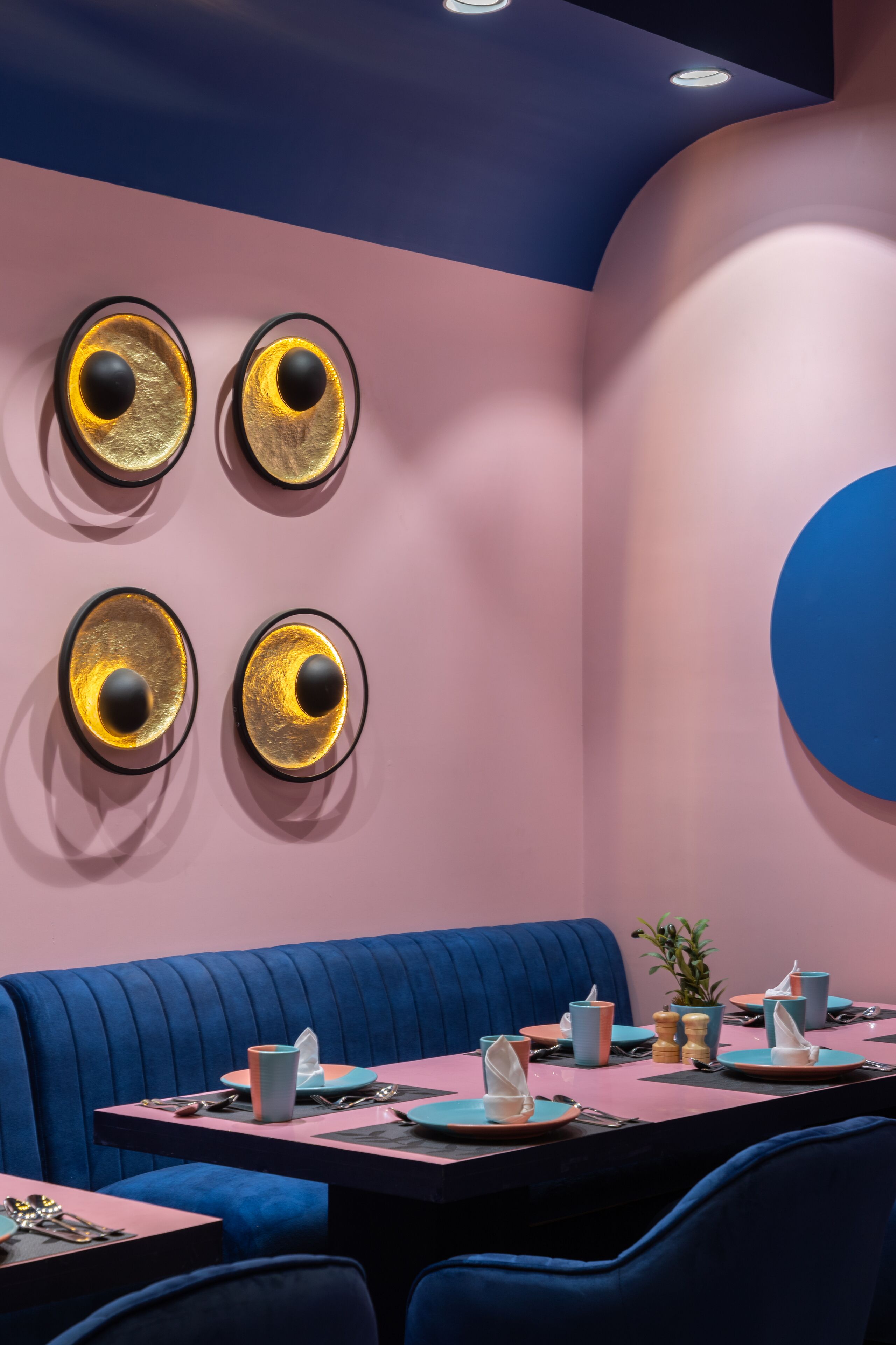 Ein modernes Restaurant mit Pink- und Blautönen, das einen Sitzbereich mit blauen Sitzecken sowie einen Tisch mit Tellern, Tassen und Besteck bietet.