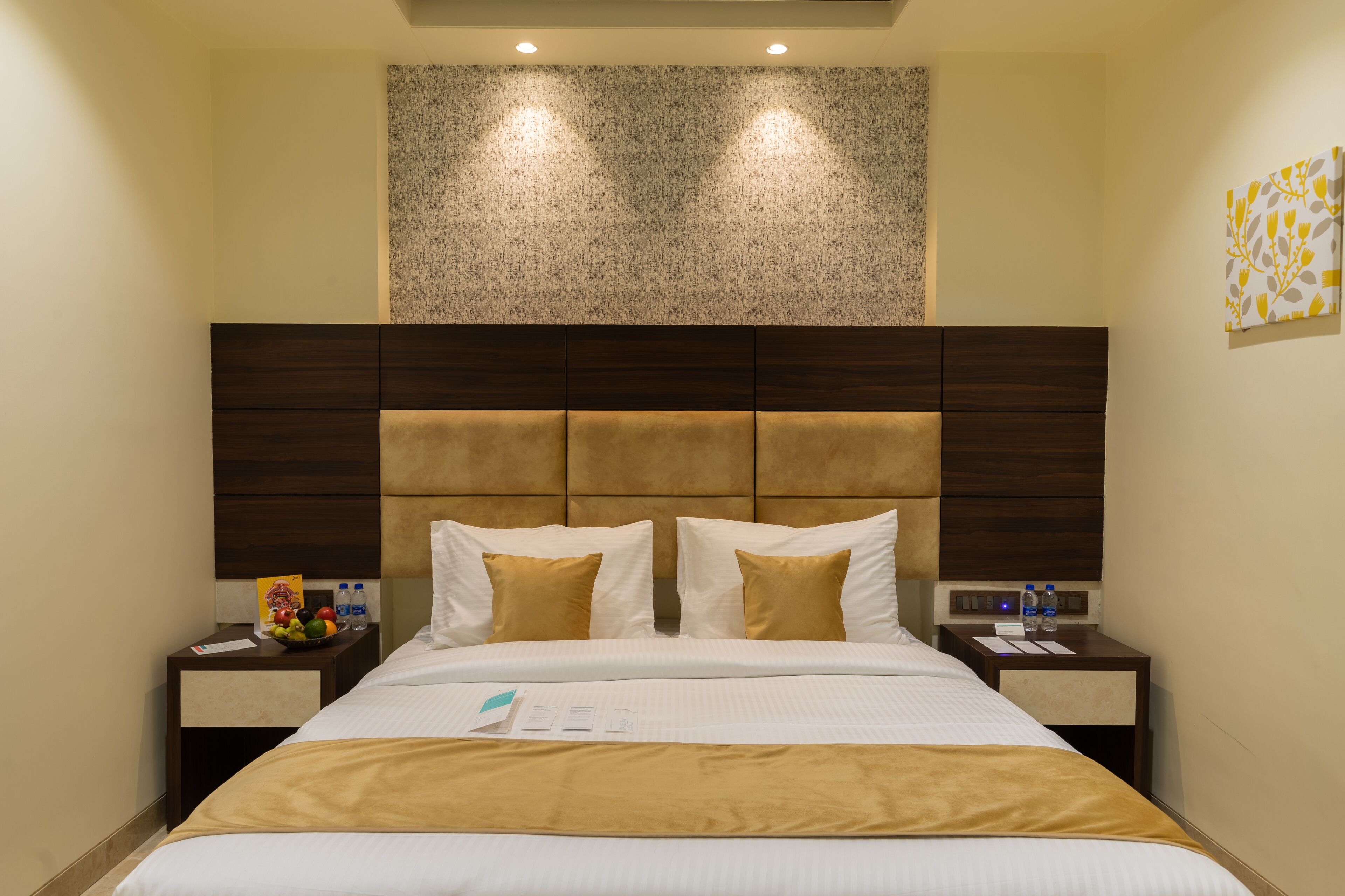 A hotel room with a large bed, a headboard with a textured panel, two bedside tables with water bottles, and a fruit bowl.