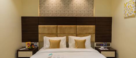 A hotel room with a large bed, a headboard with a textured panel, two bedside tables with water bottles, and a fruit bowl.