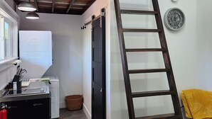 Ein Schlafzimmer im Loft-Stil mit Etagenbett, einer Leiter, einer Küchenzeile mit Spüle und Herd sowie einem kleinen Esstisch.
