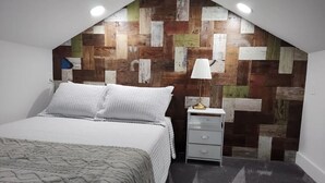 Ein Schlafzimmer mit schrägem Dach, einem Bett mit weißer Bettwäsche, einem Nachttisch mit Lampe und einer Wand mit Holzpaneelen.