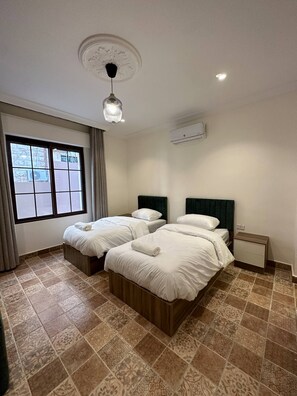 A hotel room with two beds, a window with curtains, a ceiling light, and an air conditioning unit.
