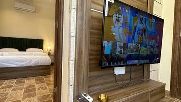 A hotel room with a large flat-screen TV mounted on a wooden wall, a bed with white linens, patterned floor tiles, and a small table with a remote control and a decorative bowl.