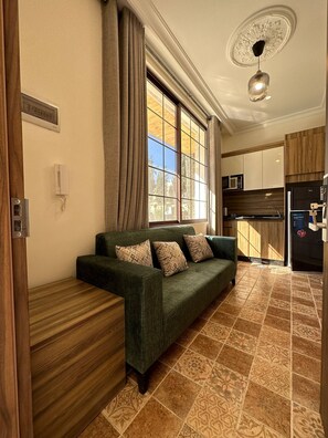 A modern living room with a sofa, patterned cushions, a wooden side table, a kitchen area with cabinets and appliances, and a large window with curtains.
