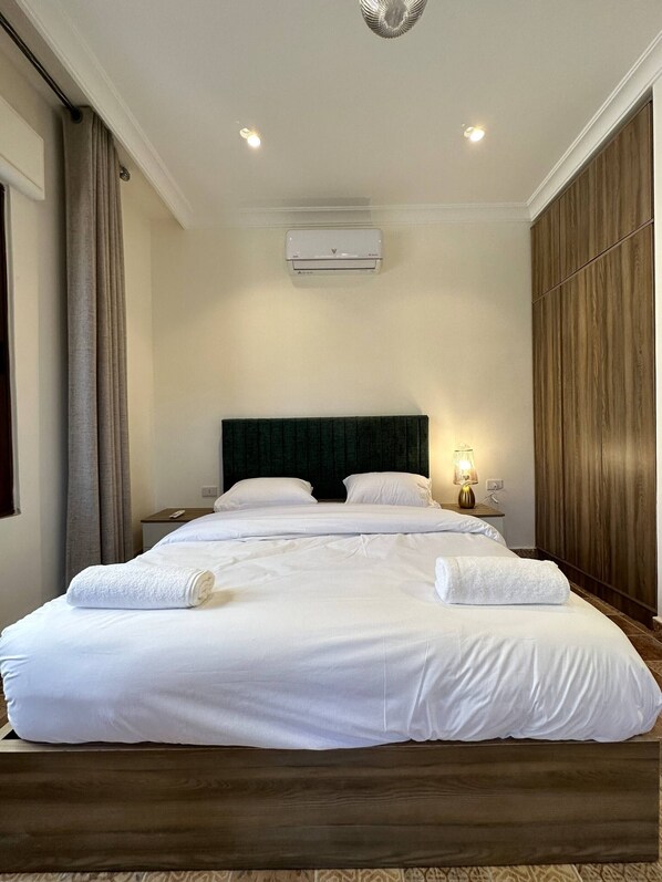 A large bed with white linens, a dark headboard, a bedside table with a lamp, and an air conditioning unit on the wall.