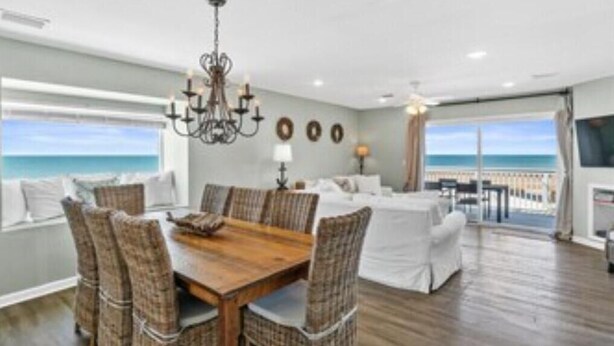 A spacious living room with a wooden dining table, wicker chairs, and a chandelier. There is a large window offering a view of the ocean.