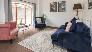 Een woonkamer met een roze fauteuil, een blauwe fauteuil, een blauwe bank, een ronde salontafel en een groot vloerkleed. Twee ingelijste foto's hangen aan de muur, een vloerlamp en een raam met gordijnen.