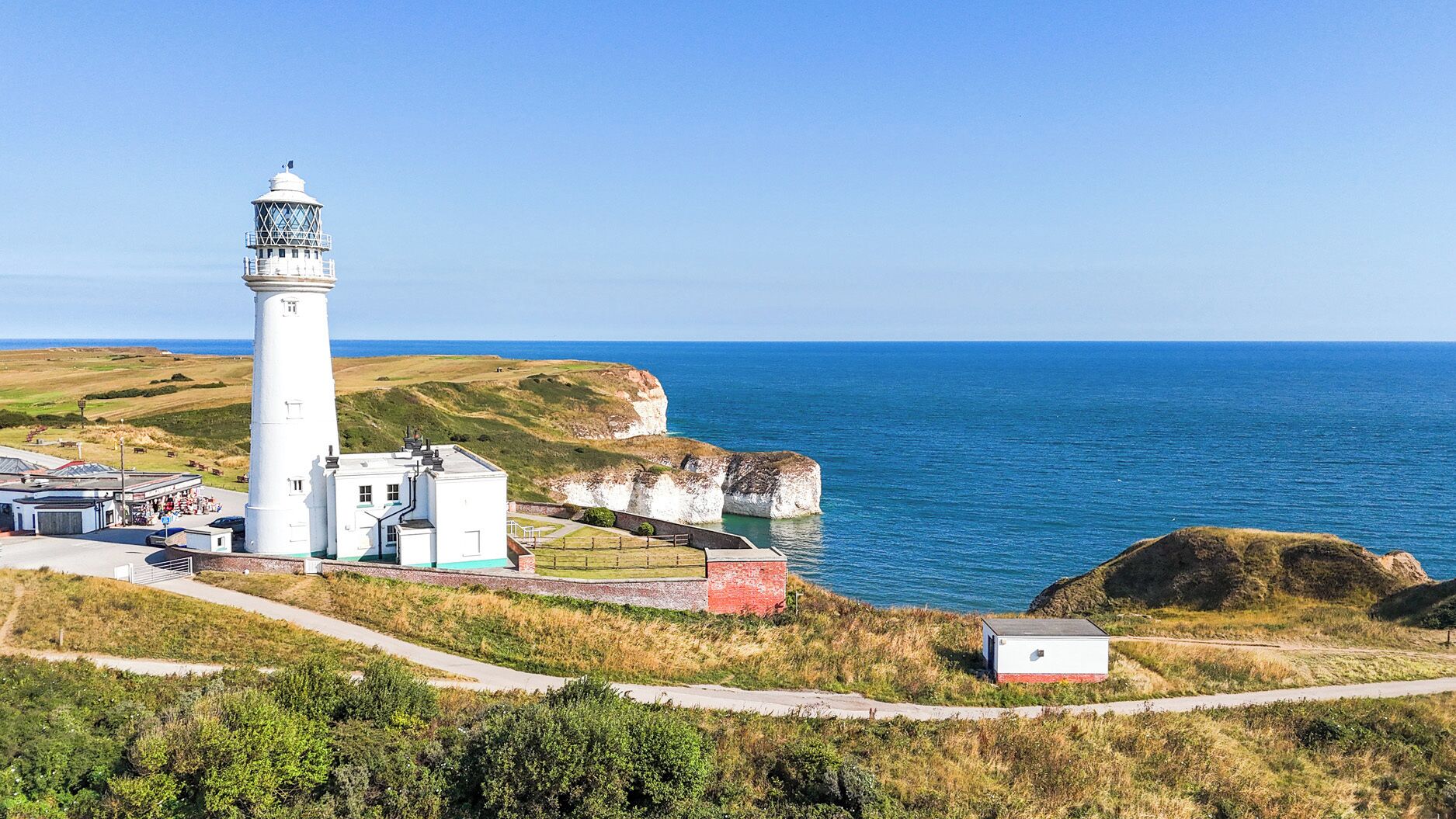 Un phare doté d’une tour blanche et d’un bâtiment plus petit attenant, le tout entouré d’un terrain herbeux et bordé par la mer.