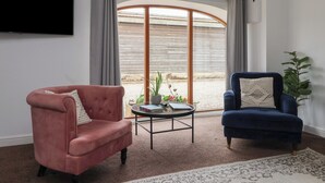 Een woonkamer met een roze fauteuil, een blauwe fauteuil, een ronde salontafel en een groot raam met gordijnen.