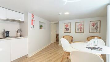 A modern kitchen with white cabinets, a coffee machine, and a small plant. There is a dining area with a white table and chairs, and framed botanical prints on the wall.