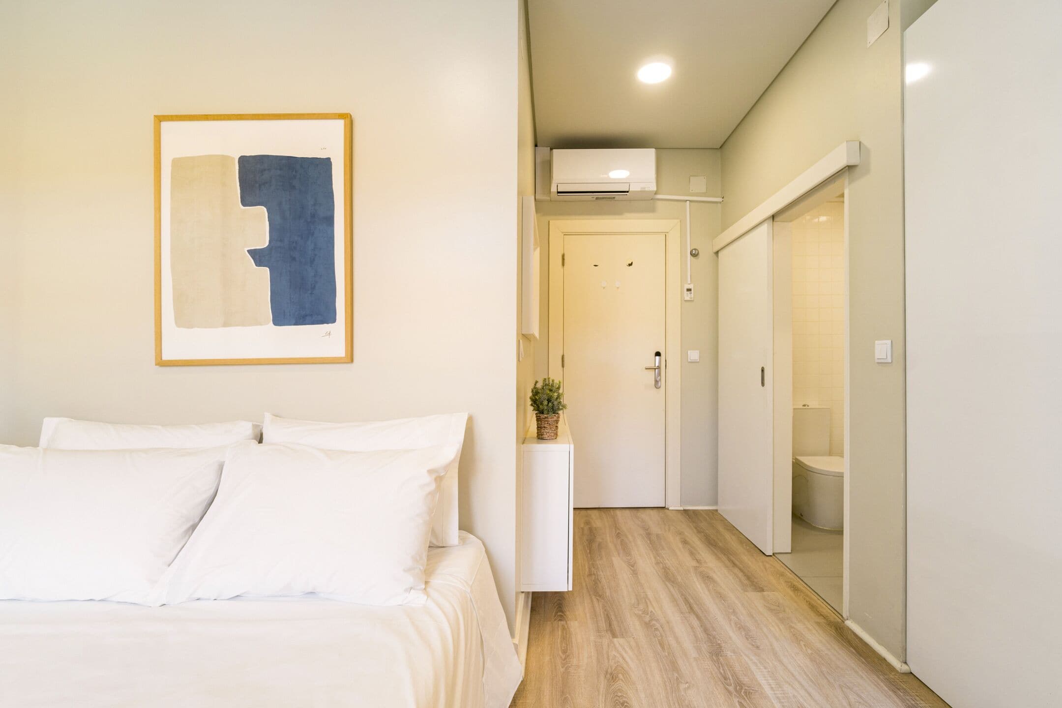 A hotel room with a bed, a wall-mounted abstract painting, a small plant on a white console table, and a hallway leading to a bathroom.