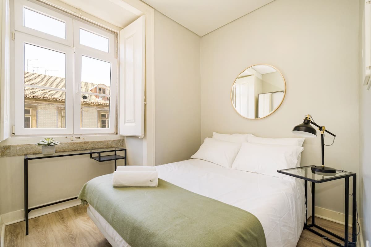 Een slaapkamer met een bed, een nachtkastje met een lamp, een ronde spiegel en een raam met uitzicht op gebouwen.