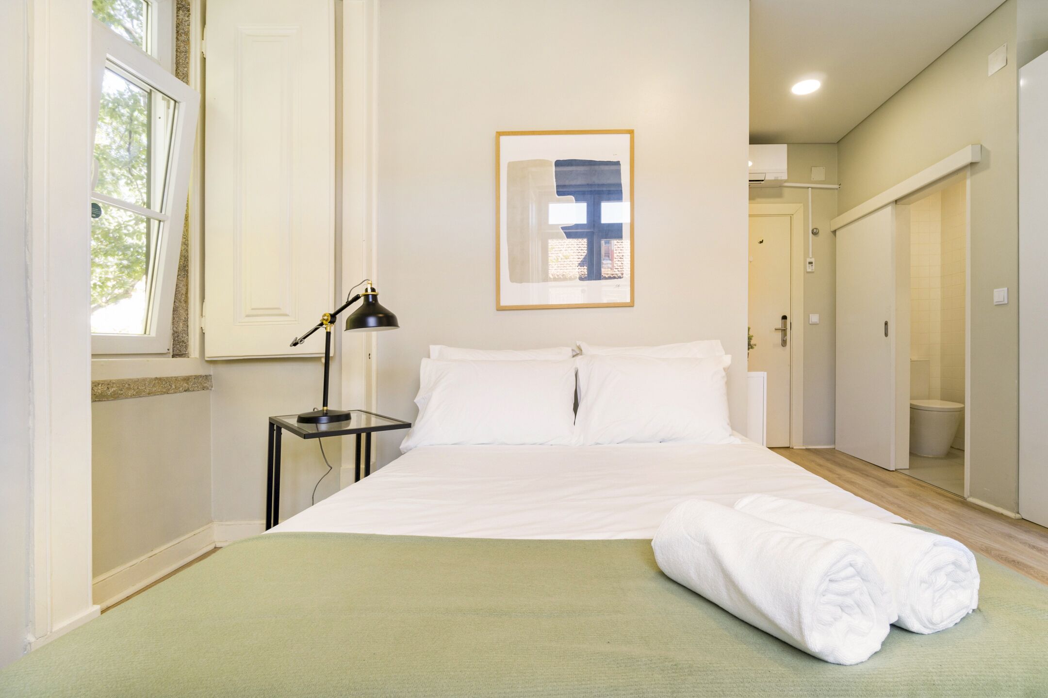 A neatly made bed with white linens and a green bedspread, a bedside lamp, and a framed picture on the wall.