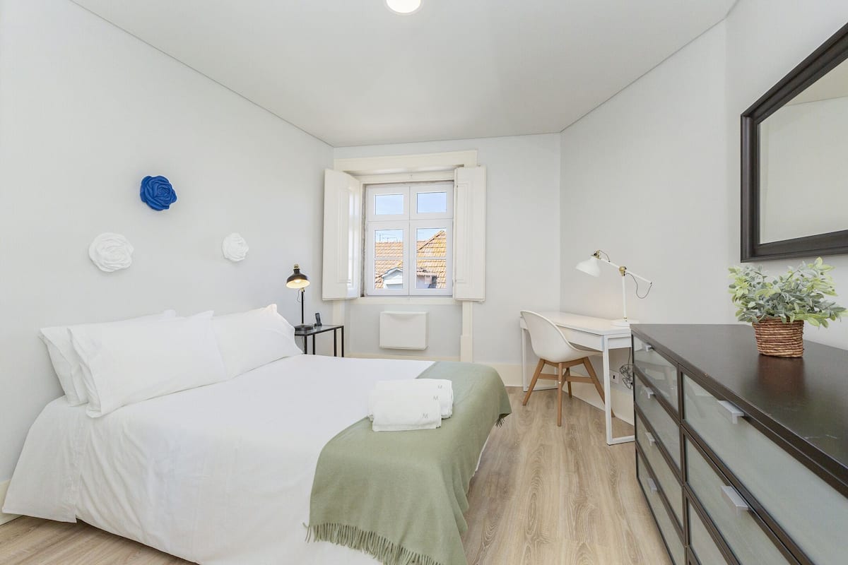 Een slaapkamer met een bed, een bureau, een stoel, een raam en een plant.