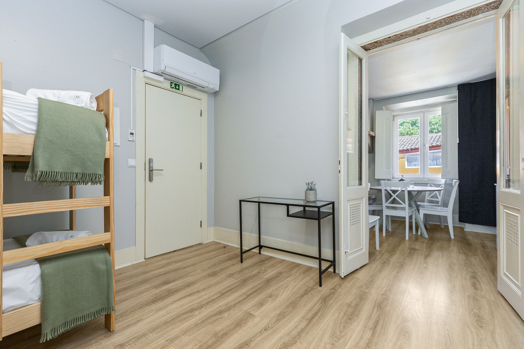 Una habitación con literas, una mesita pequeña, una puerta con letrero de salida de emergencia y vista hacia un comedor a través de una puerta abierta.