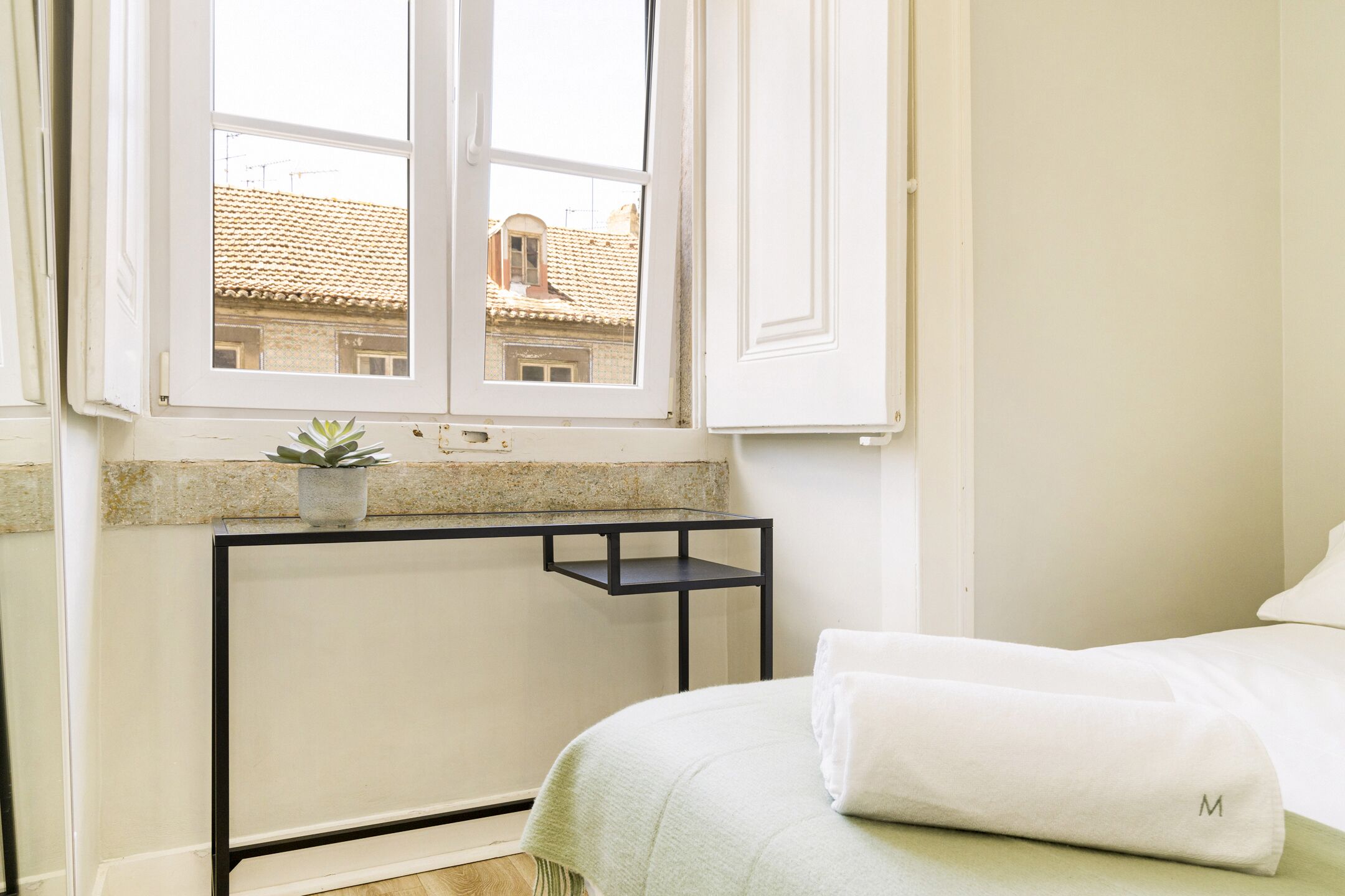 Un dormitorio con cama, ventana, una mesita con planta y vistas a edificios.
