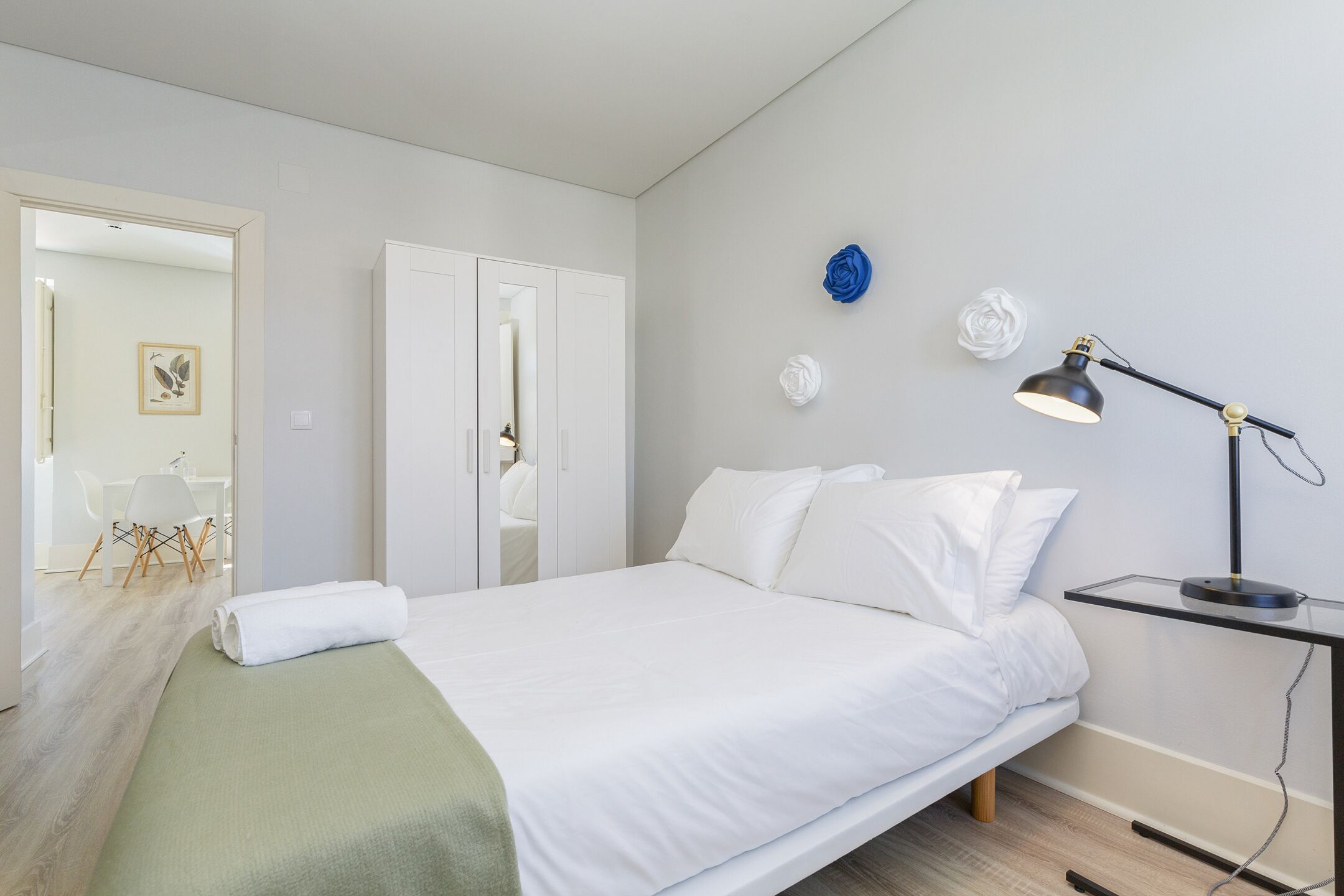 A neatly made bed with white linens, a bedside table with a lamp, and a wall-mounted flower decoration.