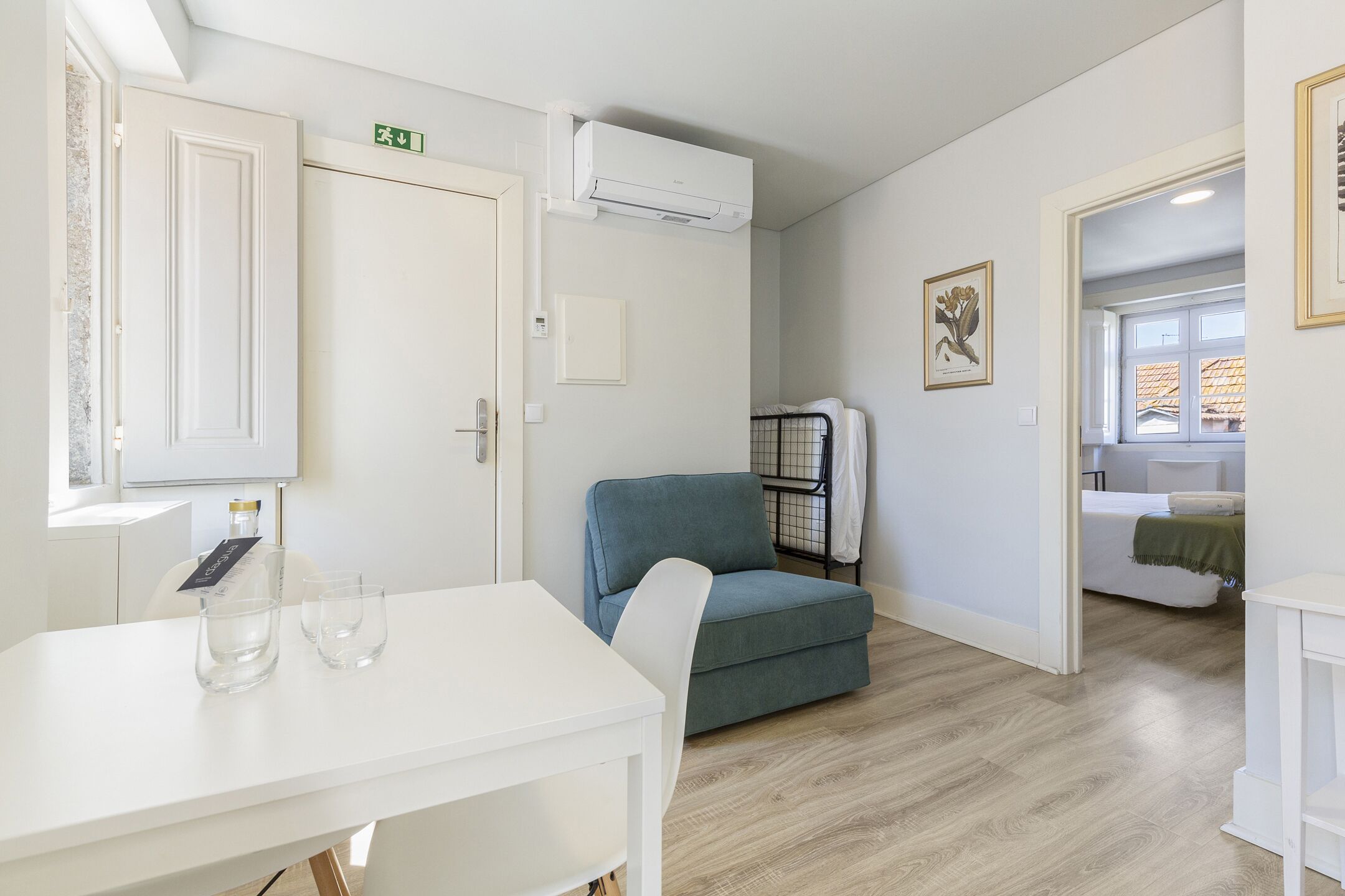 Una habitación con una mesa blanca, una silla azul, un sofá blanco y una cama con una manta verde.
