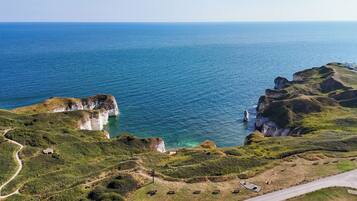 A coastal landscape with cliffs, a clear blue sea, and a paved path leading to a small building.
