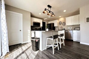 A modern kitchen with white cabinets, a central island with bar stools, stainless steel appliances, and a trash can.