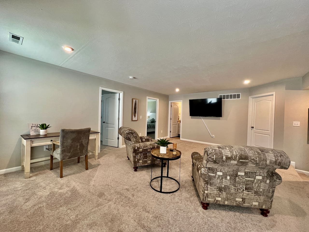 A spacious living room with a carpeted floor, a flat-screen TV mounted on the wall, and a seating area with two patterned armchairs and a sofa.