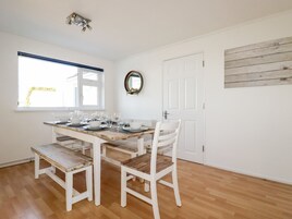 Un comedor con una mesa de madera dispuesta para cuatro personas, sillas blancas, una ventana con persianas, una puerta y un espejo fijado a la pared.