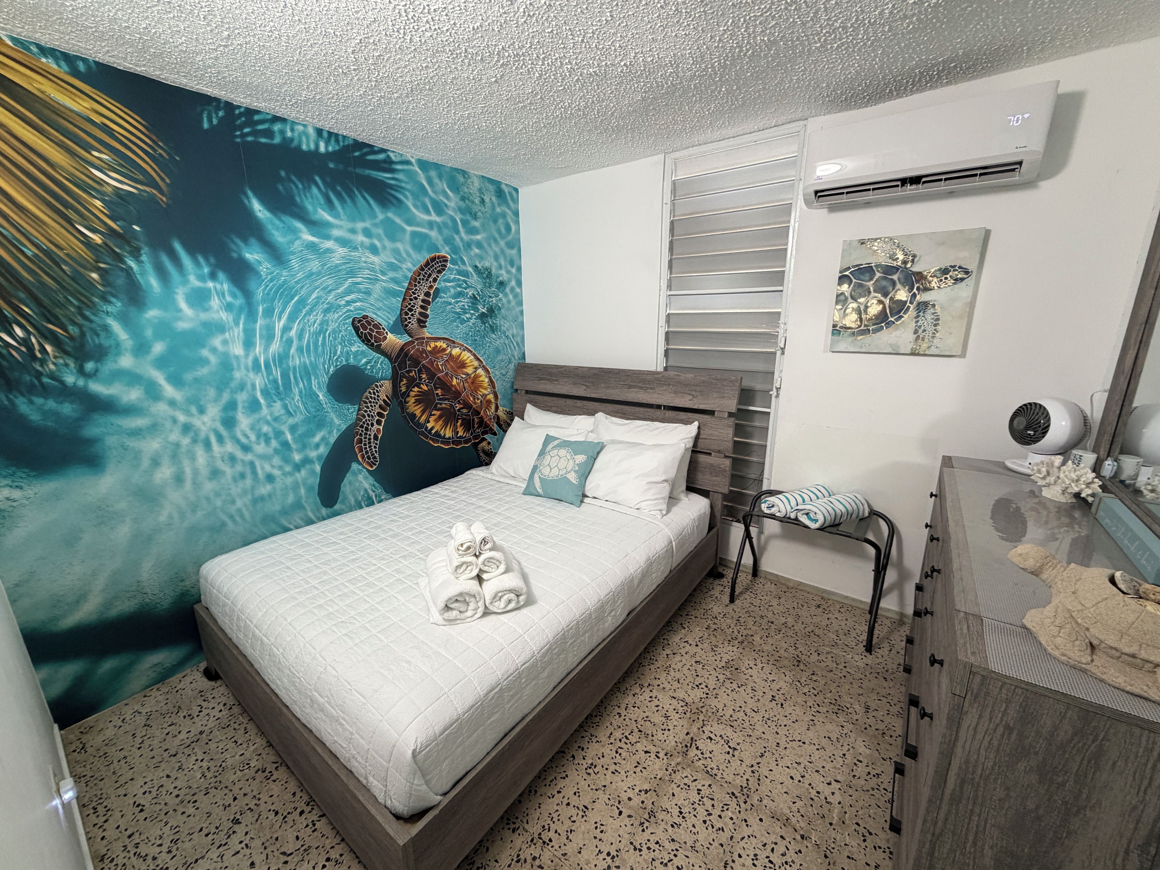 A bedroom with a bed, a wall mural of a sea turtle, an air conditioning unit, and a dresser.