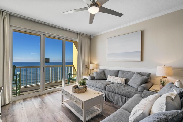 A living room with a large sliding glass door leading to a balcony with a view of the ocean. There is a ceiling fan, a sofa, a coffee table, and a lamp.