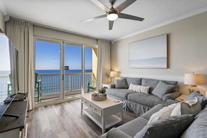 A modern living room with a sofa, a coffee table, and a TV. There is a balcony with a view of the ocean. - Calypso 2-1002 West Tower! Gulffront, Free Beach Service and Walk to Pier Park! (Panama City Beach)