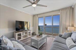 A living room with a large TV, a ceiling fan, and a view of the ocean.