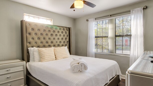A neatly made bed with a tufted headboard, a bedside table with drawers, and a window with blinds. - Savannah Home w/ Yard ~ 3 Mi to Downtown! (Savannah)