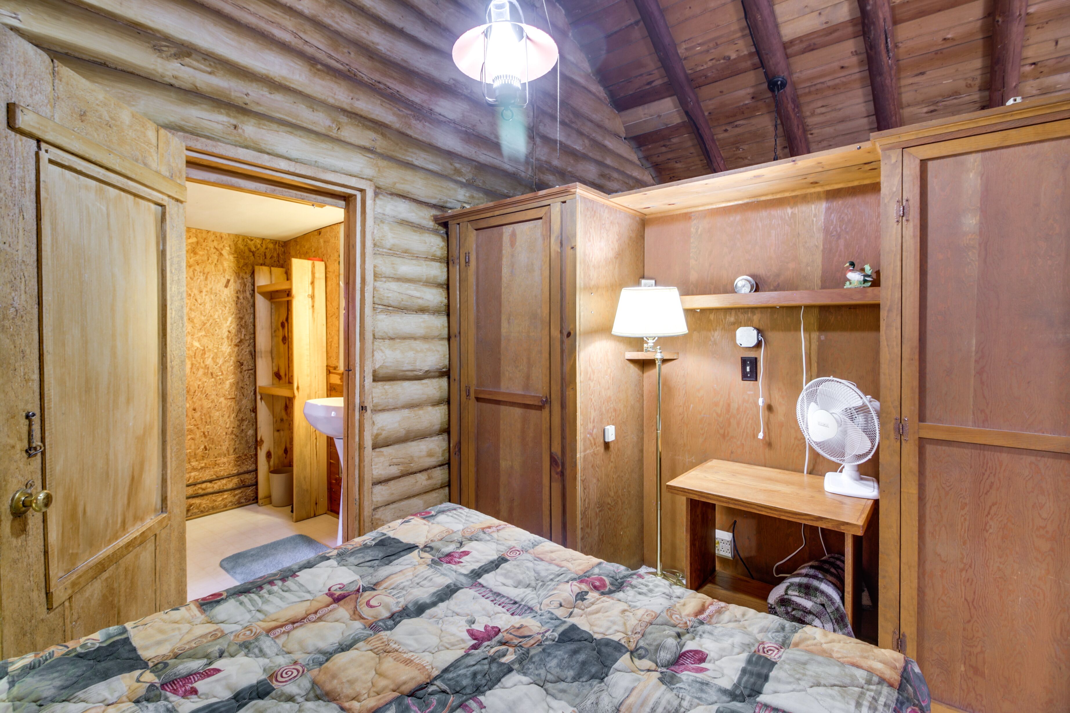 Ein Blockhauszimmer mit einem Bett, einem Holzschrank, einem Schreibtisch mit Lampe und einem Ventilator.