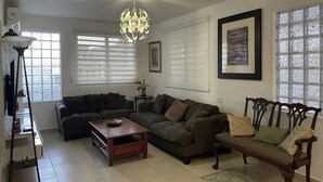 Una sala de estar con un sofá modular, una mesa de centro, una lámpara de pie y una araña. Se ven ventanas con persianas, un cuadro enmarcado en la pared y una mesita con un florero.