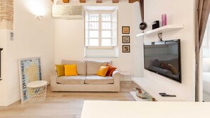 A cozy living room with a beige sofa, a flat-screen TV, and a window with white shutters. - Luxury modern Studio in historic district (Cagliari)