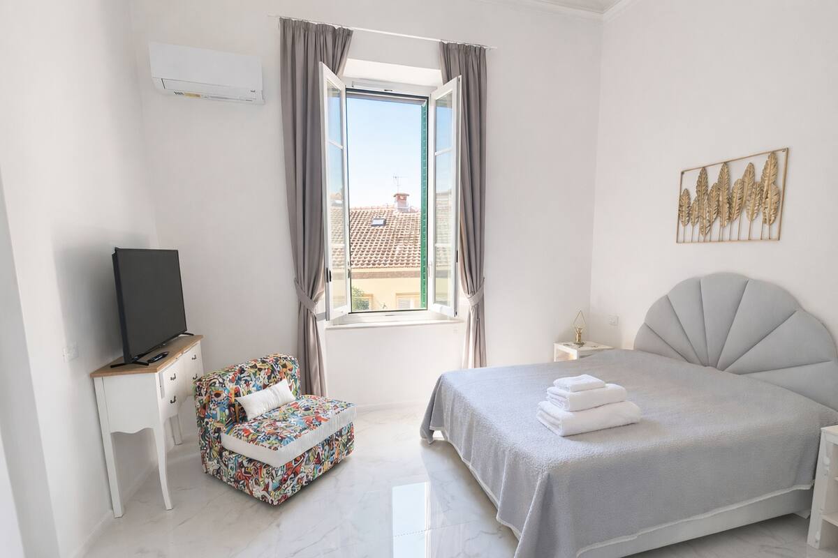 A bedroom with a bed, a TV on a stand, a window with curtains, a small table, and a chair.