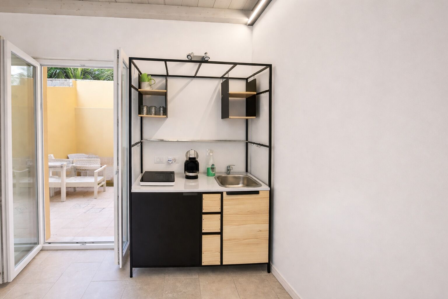 A compact kitchen with a black countertop, a sink, and a coffee machine. There is a small shelf above the countertop and a sliding door leading to a patio area with outdoor furniture.