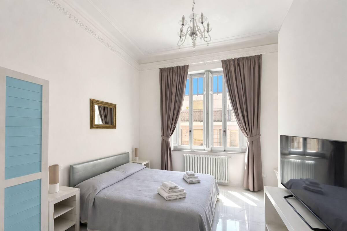 A bedroom with a bed, a TV, a window with curtains, a radiator, and a wall-mounted picture frame.