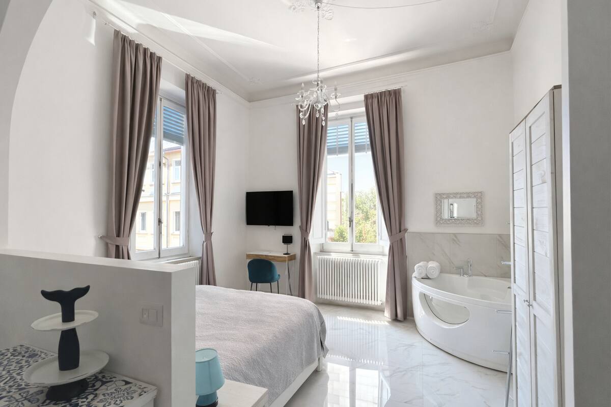 A modern hotel room with a bed, a desk, a chair, a TV, a bathroom with a bathtub, and a window with curtains.