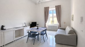 Quarto moderno e bem iluminado, com mesa de jantar, cadeiras, sofá, televisão e lustre.
