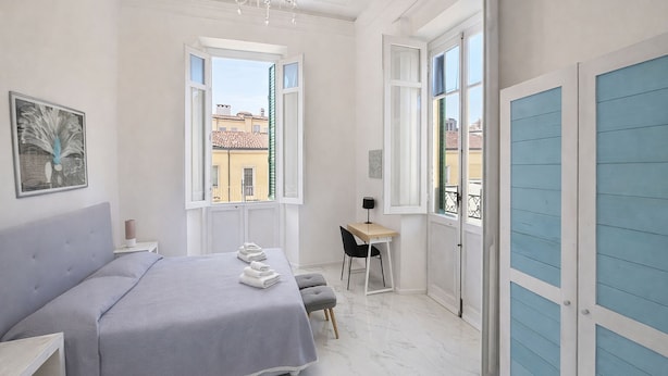 Quarto com cama, escrivaninha, cadeira, lustre e janelas amplas.