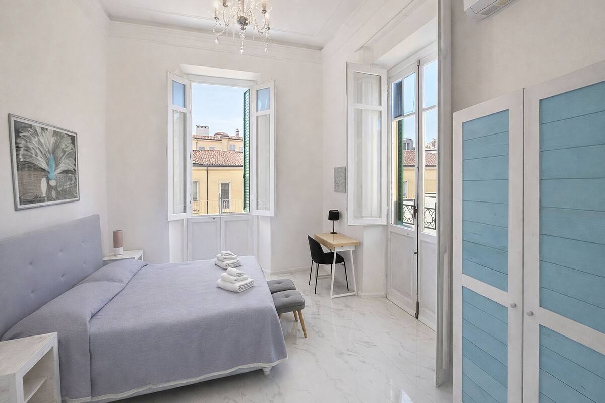 A bedroom with a bed, a desk, a chair, a chandelier, and large windows.