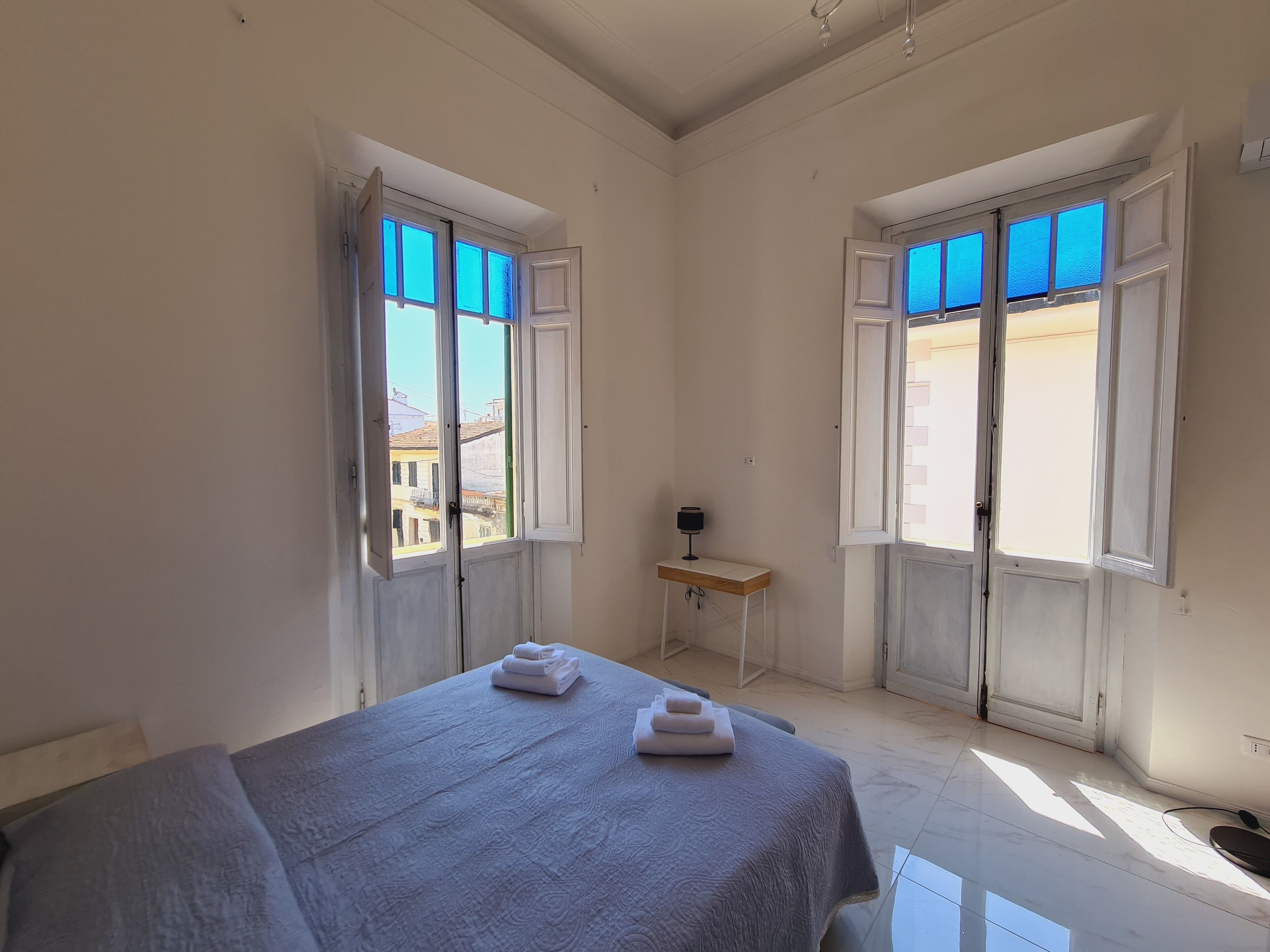 Une pièce avec un lit, deux fenêtres dont les volets sont ouverts, une petite table avec une lampe, et une vue sur les bâtiments à l’extérieur.