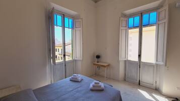 Une pièce avec un lit, deux fenêtres dont les volets sont ouverts, une petite table avec une lampe, et une vue sur les bâtiments à l’extérieur.