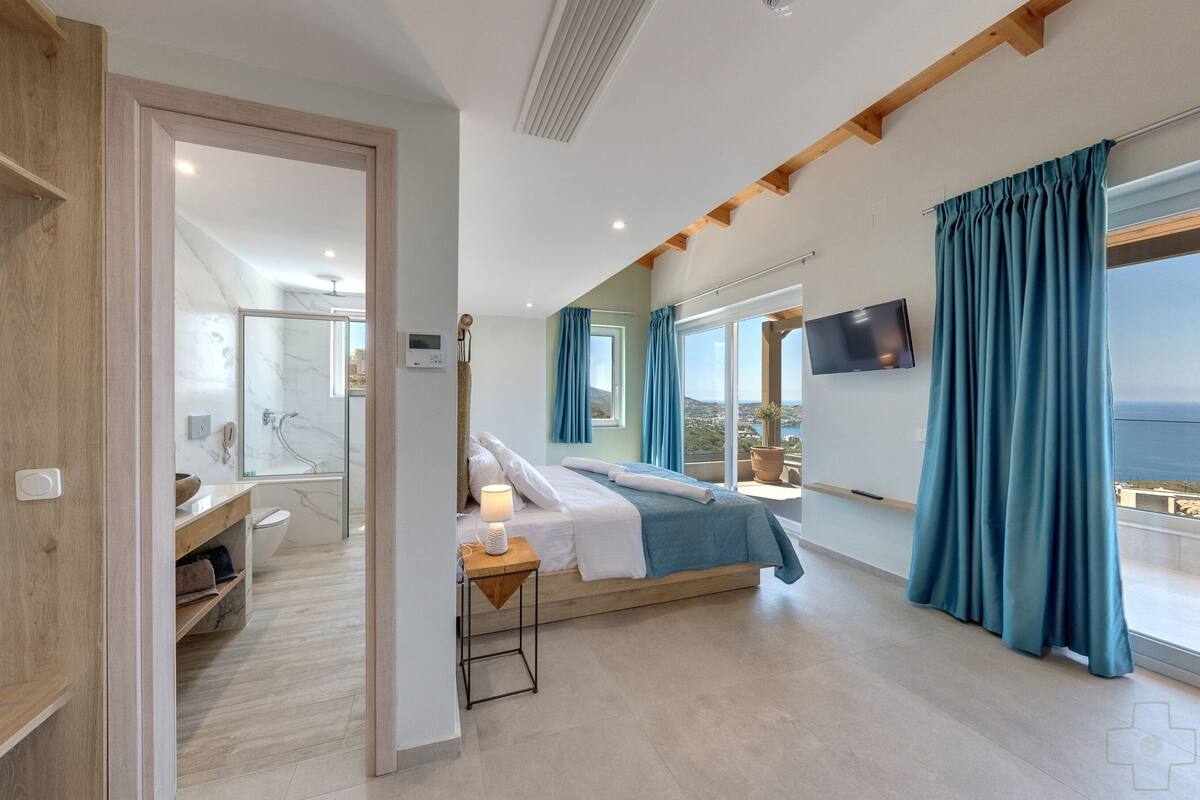 Une chambre d’hôtel moderne dotée d’un grand lit, d’une salle de bain avec une douche en verre, d’une vue sur la mer et d’un téléviseur à écran plat fixé au mur.