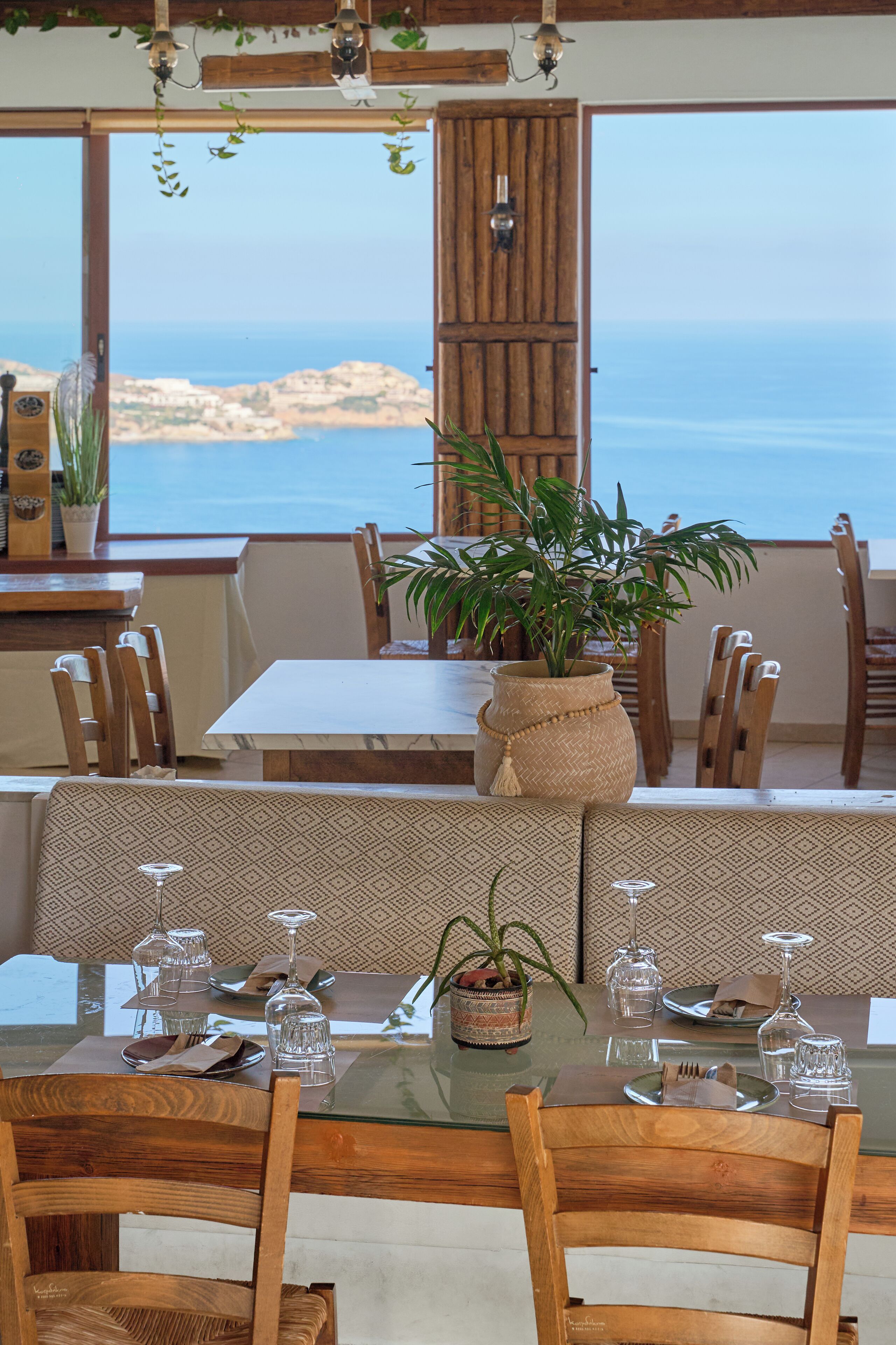 Un comedor con una mesa dispuesta para dos, sillas de madera, una planta grande en maceta y vista al mar.